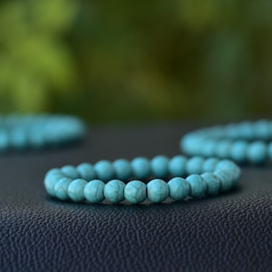May include: Three turquoise beaded bracelets. The bracelets are made of round, light blue beads with a marbled appearance. The bracelets are arranged on a dark surface with a blurred green background.