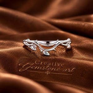 May include: A silver-toned ring featuring a delicate, curved band designed to resemble a thin, winding vine with small, sculpted leaves. The metal has a polished, reflective finish. The text Creative Gemstone art is visible below the ring.