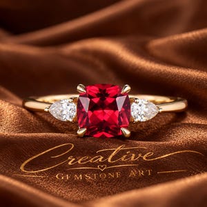 Peut inclure: Une bague en or avec une pierre rouge centrale taille coussin, encadrée par deux pierres claires taille poire. Le texte Creative GEMSTONE ART est visible sous la bague. L'anneau est en or poli et le sertissage maintient les pierres.