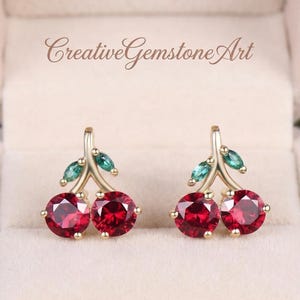 May include: Gold-toned cherry earrings with red gemstone 'cherries' and green leaf accents. The earrings are set against a soft, neutral background. The text "Creative Gemstone Art" is visible at the top.