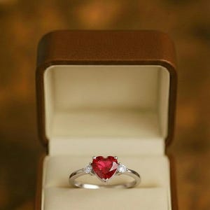Puede incluir: Un anillo de plata con una piedra preciosa roja en forma de corazón, flanqueada por dos piedras preciosas claras, redondas y más pequeñas. El anillo se presenta en un joyero abierto, marrón, con aspecto de cuero y un interior color crema. El fondo está borroso.