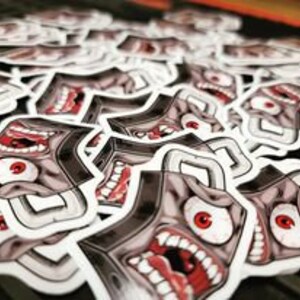 May include: A sticker featuring a cartoon monster with a large mouth, one eye, and a single tooth. The monster is gray with red accents and is set against a white background.