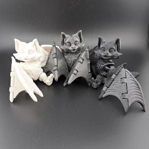 May include: Three bat figurines, one white and two black, with articulated wings. The white bat is sitting up with its wings spread out. The black bats are sitting with their wings folded behind them.