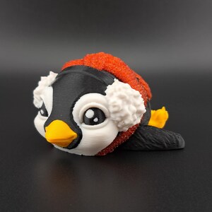 May include: A 3D printed penguin figurine wearing a red and white winter hat with earmuffs. The penguin has black and white feathers and a yellow beak.