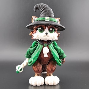 May include: A whimsical figurine of a cat dressed as a wizard. The cat wears a black witch hat with a green band, a green cape, and holds a white staff with a green tip. The cat has brown and white fur and large green eyes.