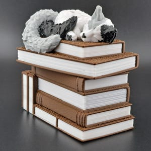 May include: A white and gray cat figurine sleeping on top of a stack of five brown books with white pages.