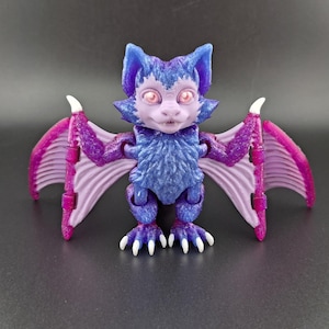 May include: A purple and pink bat-like creature with articulated wings and glowing pink eyes. The toy is made of a textured plastic and has a fuzzy appearance.