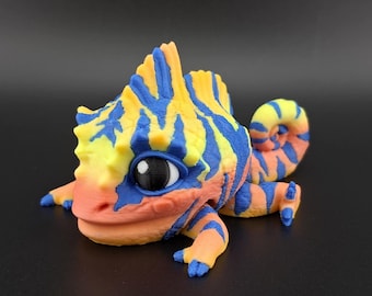 May include: A 3D printed chameleon figurine with a bright orange, yellow, and blue striped pattern. The chameleon has large, black eyes and a curled tail.