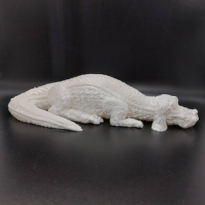 May include: A white ceramic sculpture of a crocodile lying on its belly with its head turned to the side. The crocodile has a textured surface and a realistic appearance.