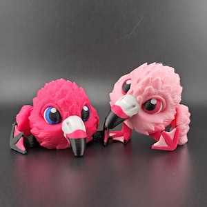 May include: Two pink 3D printed flamingo figurines with black beaks and blue eyes. The figurines are in a playful pose, with one standing and the other lying down.