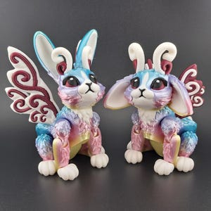 May include: Two small, colorful, 3D printed rabbit figurines with intricate details. One rabbit has wings with a burgundy swirl design. Both rabbits have white ears and blue, pink, and purple fur.