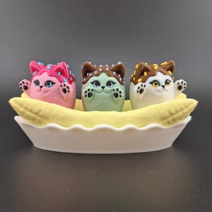 May include: Three small, colorful, cat-shaped figurines are sitting in a banana-shaped boat. The cats are decorated with chocolate, sprinkles, and frosting. The boat is white and has a scalloped edge.