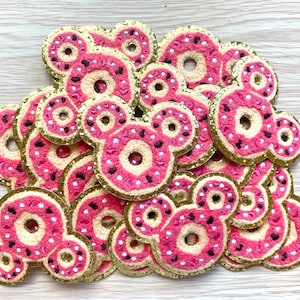 May include: A pile of pink and gold glitter donut-shaped felt patches. The donuts have a white center and pink frosting with sprinkles.