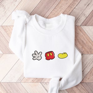 K&ouml;nnte beinhalten: Wei&szlig;er Pullover mit einem roten, gelben und wei&szlig;en Mickey Mouse-Icon-Motiv. Die Grafik enth&auml;lt Mickey Mouse's Handschuh, Shorts und Schuh.