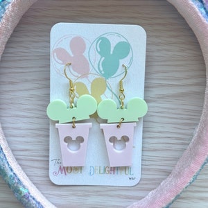 May include: A pair of pink and green dangle earrings featuring a Mickey Mouse head silhouette. The earrings are on a white card with the text "The Most Delightful Way".