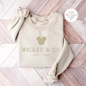 Op de afbeelding: Een beige sweatshirt met lange mouwen en ronde hals, voorzien van een geborduurd silhouet van een Mickey Mouse-hoofd boven de tekst MICKEY & CO. en EST. 1928 in een lichtbruine kleur. Een kleine cirkelvormige afbeelding rechtsboven toont de tekst Available in MULTIPLE COLORS FALL.