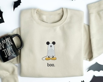 May include: A beige crewneck sweatshirt featuring an embroidered graphic of a ghost wearing round black ears and yellow shoes, with the text "boo." below it. Beside the sweatshirt is a black ceramic mug with the text "VAMPIRE JUICE" in white.