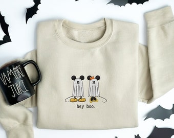 May include: A beige crewneck sweatshirt featuring an embroidered graphic of two ghost characters with mouse ears, one wearing an orange bow, and the text "hey boo." A black mug with the white text "VAMPIRE JUICE" sits beside the sweatshirt.