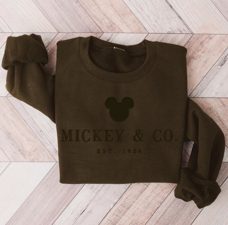 Op de afbeelding: Een donkerbruine sweater met ronde hals en een zwarte geborduurde silhouet van Mickey Mouse-oren boven de tekst MICKEY & CO. en de kleinere tekst EST. 1928. Het kledingstuk heeft lange mouwen met geribbelde boorden en een geribbelde halslijn.