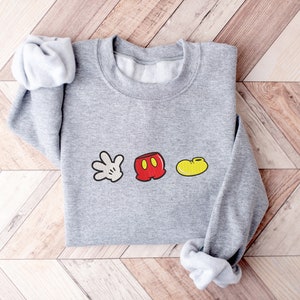 K&ouml;nnte beinhalten: Ein grauer Pullover mit einem wei&szlig;en Handschuh, einer roten Hose und einem gelben Schuh als Grafik. Die Grafik ist ein einfaches Cartoon-Design von Mickey Mouses ikonischen Merkmalen.