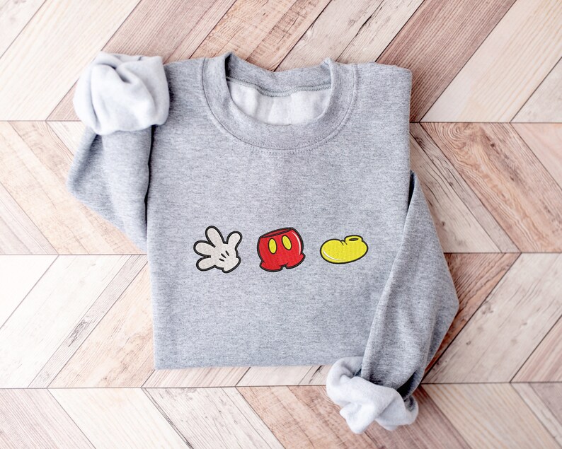 K&ouml;nnte beinhalten: Ein grauer Pullover mit einem wei&szlig;en Handschuh, einer roten Hose und einem gelben Schuh als Grafik. Die Grafik ist ein einfaches Cartoon-Design von Mickey Mouses ikonischen Merkmalen.