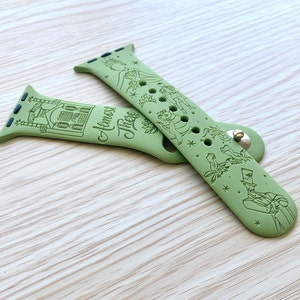 May include: A sage green silicone watch band featuring engraved illustrations of a building, a crown, a frog, and a man in a top hat, with the text "Almost There" printed in a cursive script. The band includes a metal pin closure and attachment lugs.