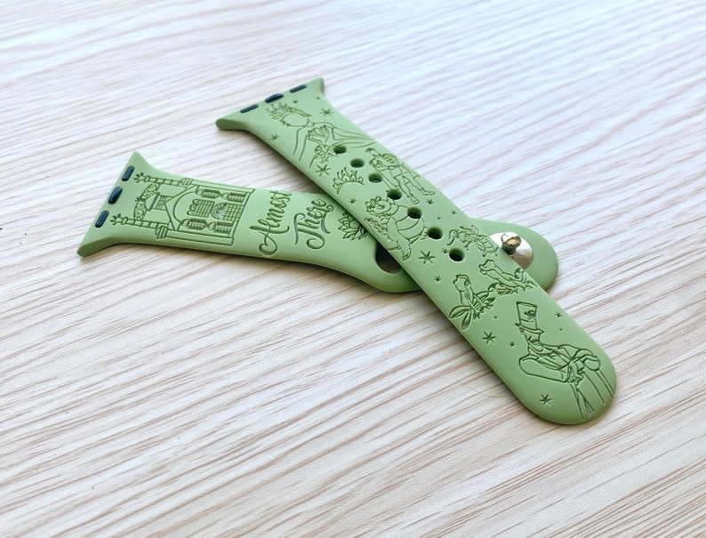May include: A sage green silicone watch band featuring engraved illustrations of a building, a crown, a frog, and a man in a top hat, with the text "Almost There" printed in a cursive script. The band includes a metal pin closure and attachment lugs.