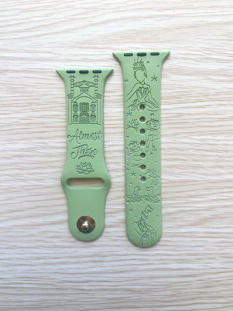 May include: A sage green silicone watch band featuring engraved illustrations of a building, a lotus flower, and the text "Almost There" on one side, with characters, a crown, and small stars engraved on the other side, including a metal pin closure.