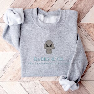 May include: Gray sweatshirt with a cartoon image of Hades, the Greek god of the underworld, wearing sunglasses. The text "Hades & Co. The Underworld Since Forever" is printed below the image.