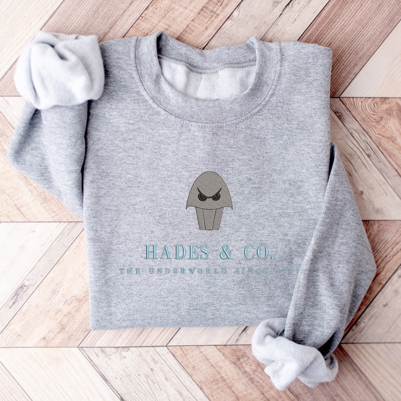 May include: Gray sweatshirt with a cartoon image of Hades, the Greek god of the underworld, wearing sunglasses. The text "Hades & Co. The Underworld Since Forever" is printed below the image.