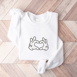 K&ouml;nnte beinhalten: Wei&szlig;er Pullover mit einer schwarzen Grafik von zwei H&auml;nden, die ein Herz bilden.