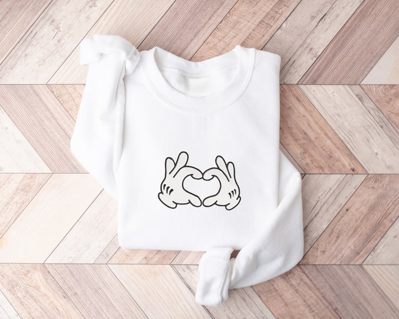 K&ouml;nnte beinhalten: Wei&szlig;er Pullover mit einer schwarzen Grafik von zwei H&auml;nden, die ein Herz bilden.