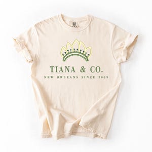 May include: Cream-colored t-shirt with rolled sleeves. The shirt features a green and yellow crown design above the text "TIANA & CO. NEW ORLEANS SINCE 2009" in green. The shirt is made of a soft, comfortable material.