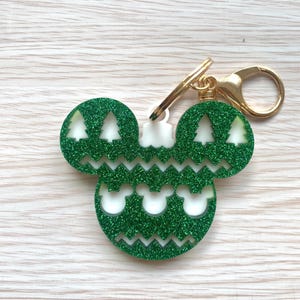 May include: A green glitter keychain shaped like a mouse head silhouette with a white base. The design features cutouts of four pine trees on the ears and three small mouse head shapes on the bottom, accented by zigzag lines and a gold clasp.