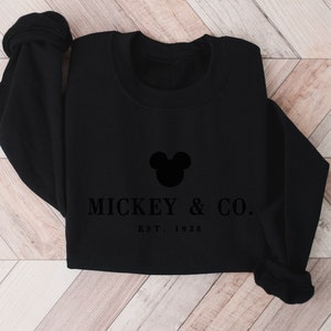 Op de afbeelding: Een zwart sweatshirt met ronde hals voorzien van een zwart silhouet van Mickey Mouse-oren, gecentreerd boven de tekst MICKEY & CO. en de tekst EST. 1928 in een schreeflettertype. Het kledingstuk heeft lange mouwen met geribbelde boorden.