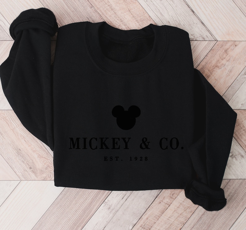 Op de afbeelding: Een zwart sweatshirt met ronde hals voorzien van een zwart silhouet van Mickey Mouse-oren, gecentreerd boven de tekst MICKEY & CO. en de tekst EST. 1928 in een schreeflettertype. Het kledingstuk heeft lange mouwen met geribbelde boorden.
