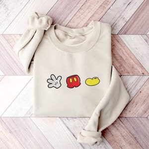 K&ouml;nnte beinhalten: Ein beiger Pullover mit einem wei&szlig;en Handschuh, einer roten Hose und einem gelben Schuh, alle mit schwarzen Umrissen, in einer Reihe auf der Vorderseite. Der Pullover ist gefaltet und auf einer Holzoberfl&auml;che abgelegt.