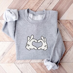 K&ouml;nnte beinhalten: Grauer Pullover mit einem wei&szlig;en Aufdruck von zwei H&auml;nden, die ein Herz bilden.