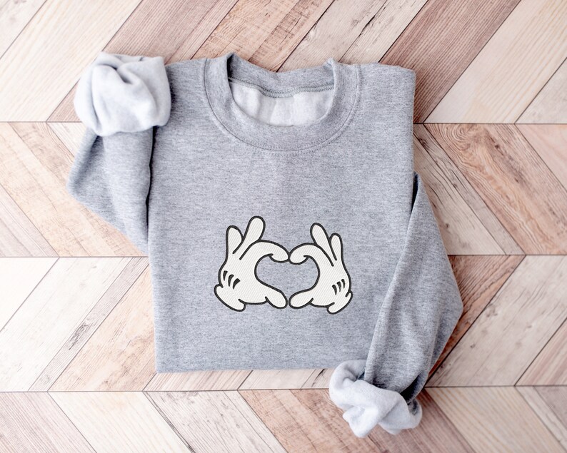 K&ouml;nnte beinhalten: Grauer Pullover mit einem wei&szlig;en Aufdruck von zwei H&auml;nden, die ein Herz bilden.