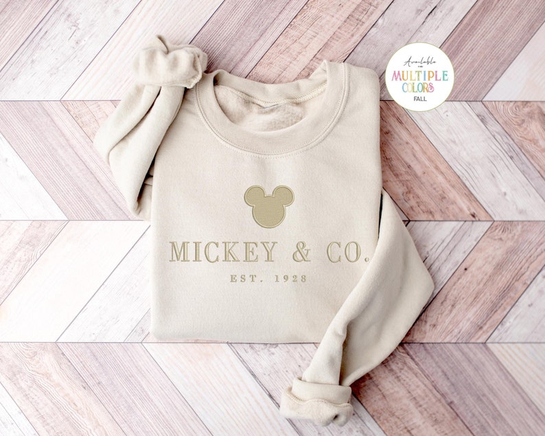 Op de afbeelding: Een beige sweatshirt met lange mouwen en ronde hals, voorzien van een geborduurd silhouet van een Mickey Mouse-hoofd boven de tekst MICKEY & CO. en EST. 1928 in een lichtbruine kleur. Een kleine cirkelvormige afbeelding rechtsboven toont de tekst Available in MULTIPLE COLORS FALL.