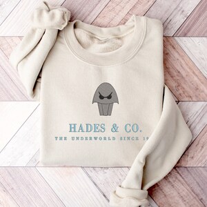 May include: A beige sweatshirt with a gray cartoon image of a creature with a single eye and the text "Hades & Co. The Underworld Since 19..."
