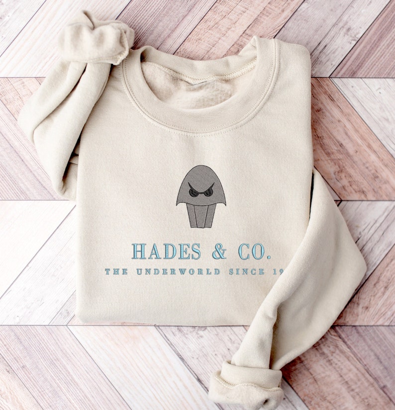 May include: A beige sweatshirt with a gray cartoon image of a creature with a single eye and the text "Hades & Co. The Underworld Since 19..."