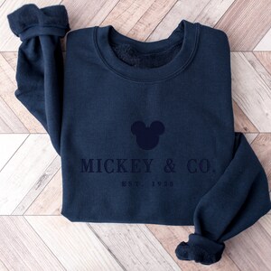 Op de afbeelding: Een marineblauw sweatshirt met ronde hals en een donkerblauw geborduurde Mickey Mouse-hoofdsilhouet boven de tekst MICKEY & CO. en EST. 1928. Het kledingstuk heeft lange mouwen met geribbelde boorden en een zachte fleece voering bij de kraag.