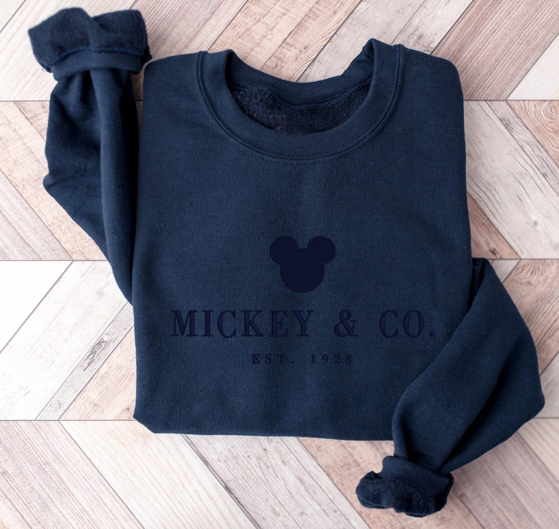 Op de afbeelding: Een marineblauw sweatshirt met ronde hals en een donkerblauw geborduurde Mickey Mouse-hoofdsilhouet boven de tekst MICKEY & CO. en EST. 1928. Het kledingstuk heeft lange mouwen met geribbelde boorden en een zachte fleece voering bij de kraag.