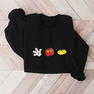 K&ouml;nnte beinhalten: Schwarzer Pullover mit einem wei&szlig;en Handschuh, einer roten Hose und einem gelben Schuh, die auf der Vorderseite gestickt sind. Die gestickten Bilder sind im Stil von Mickey Mouse.