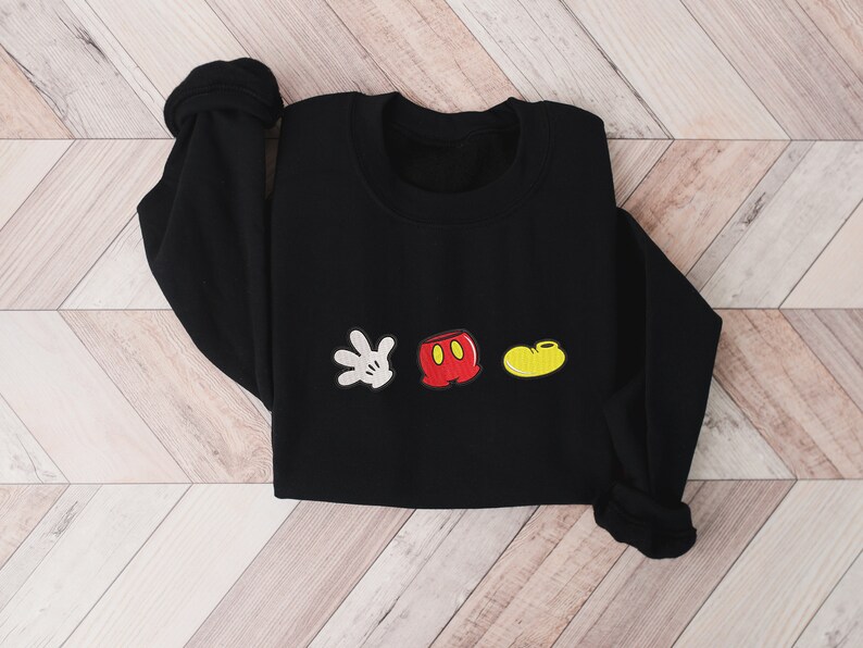K&ouml;nnte beinhalten: Schwarzer Pullover mit einem wei&szlig;en Handschuh, einer roten Hose und einem gelben Schuh, die auf der Vorderseite gestickt sind. Die gestickten Bilder sind im Stil von Mickey Mouse.