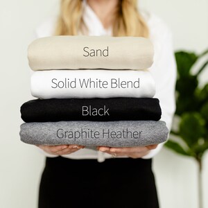 May include: A stack of folded t-shirts in four colors: sand, solid white blend, black, and graphite heather.