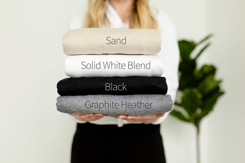 May include: A stack of folded t-shirts in four colors: sand, solid white blend, black, and graphite heather.