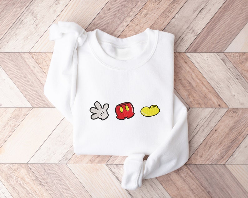 K&ouml;nnte beinhalten: Wei&szlig;er Pullover mit einem roten, gelben und wei&szlig;en Mickey Mouse-Icon-Motiv. Die Grafik enth&auml;lt Mickey Mouse's Handschuh, Shorts und Schuh.