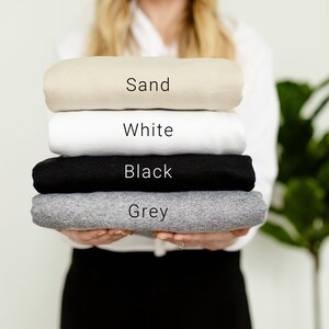 K&ouml;nnte beinhalten: Ein Stapel gefalteter T-Shirts in verschiedenen Farben: Sand, Wei&szlig;, Schwarz und Grau.